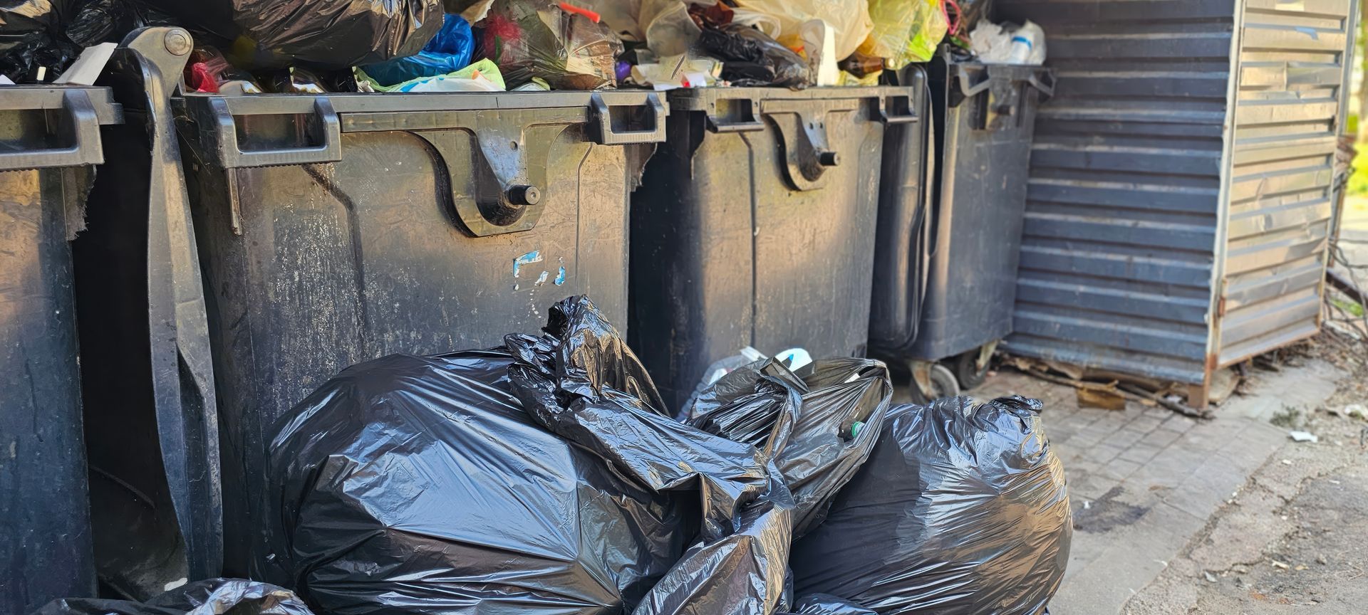 Des sacs-poubelles noirs empilés à côté de grands conteneurs à ordures dans une ruelle
