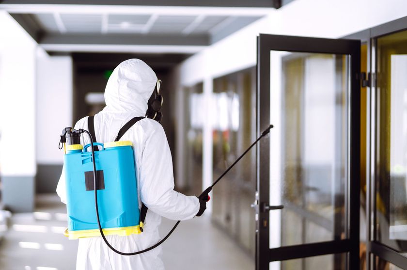 Un ouvrier en combinaison de protection blanche pulvérise du désinfectant dans un couloir dont la porte est ouverte.