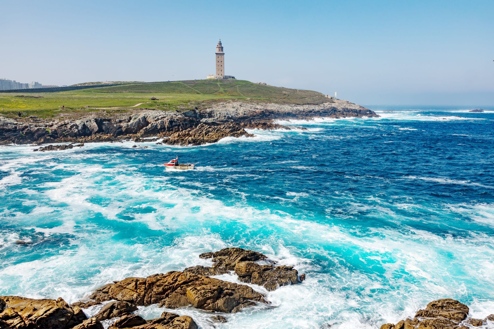 Cala rocosa junto al mar con olas turquesas, un kayak rojo y un faro en el promontorio.