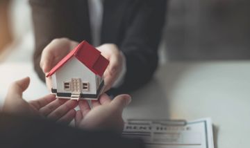 Manos intercambiando una pequeña maqueta de una casa con techo rojo sobre papeles en un escritorio.