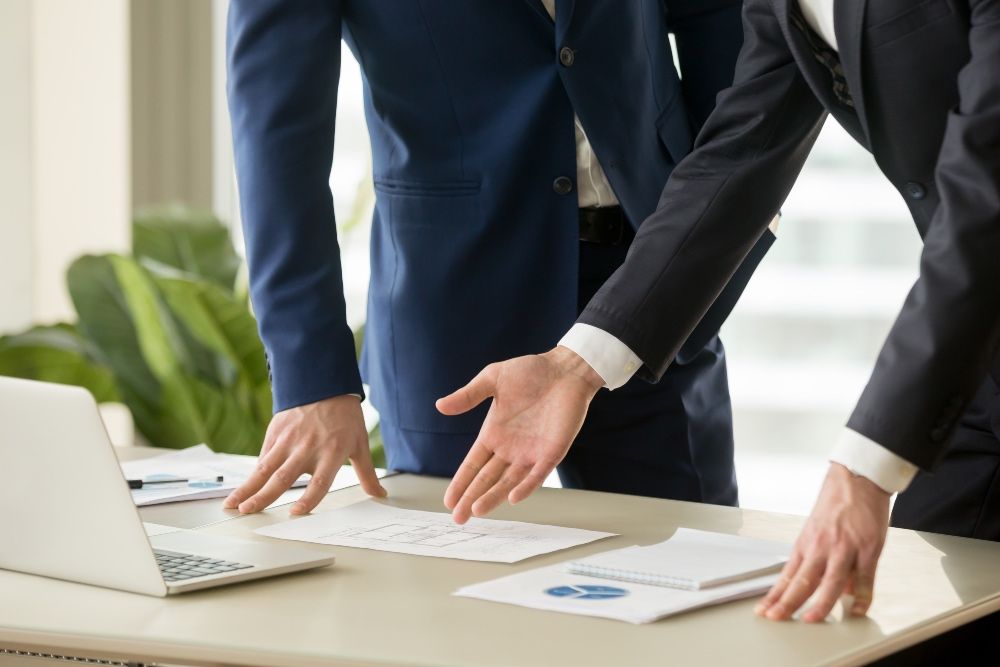 Dos hombres de negocios con traje señalan unos documentos que hay sobre un escritorio junto a un ordenador portátil y una taza de café.