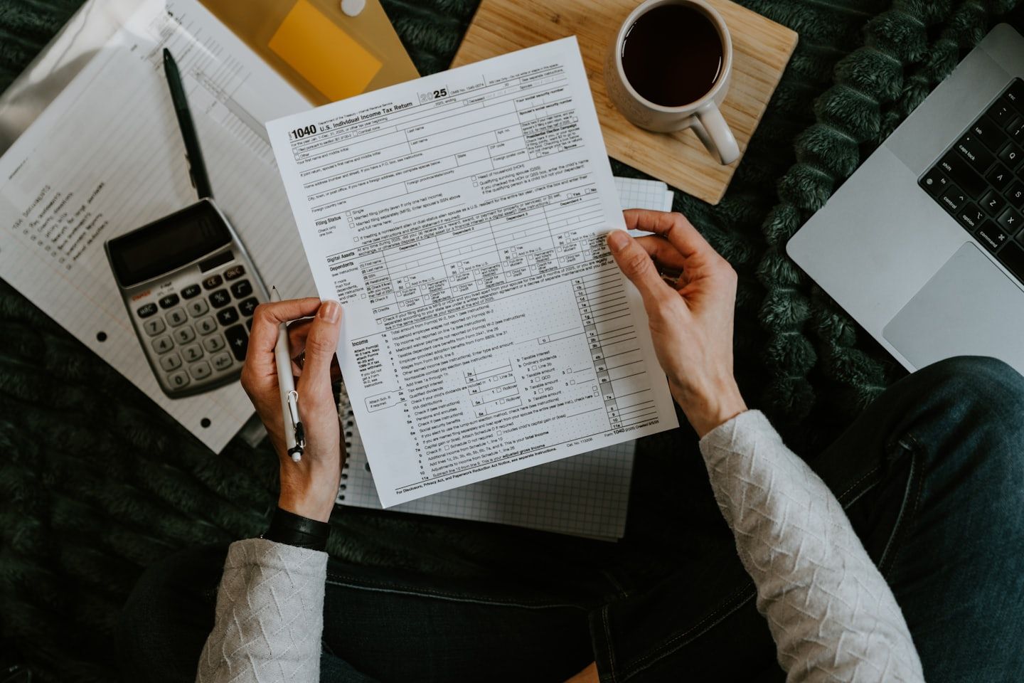 Manos revisando documentos financieros en un escritorio con calculadora, café y computadora portátil.