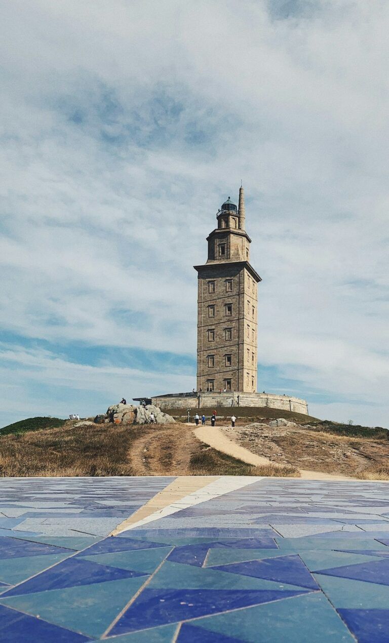 Una alta torre con aspecto de faro se alza sobre una colina rocosa junto a un paseo marítimo cubierto de mosaicos