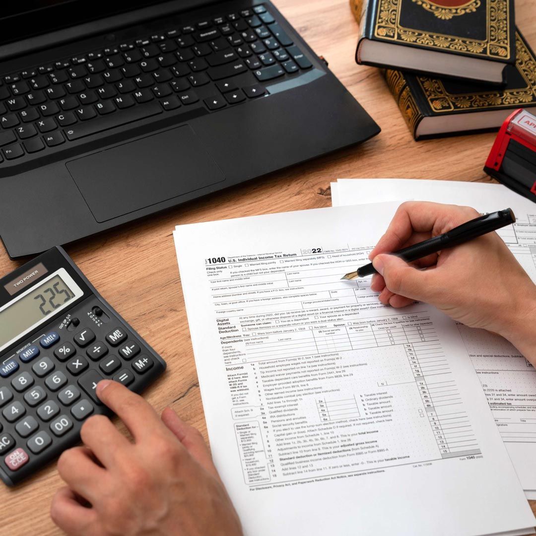 Manos calculando en una calculadora mientras escribe en documentos financieros junto a una computadora portátil y libros.