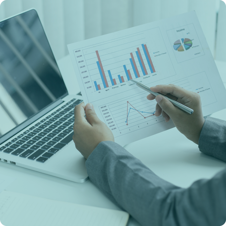 A person is holding a piece of paper with a graph on it in front of a laptop computer.