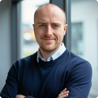 Man with light skin, blue sweater, and collared shirt smiling indoors.