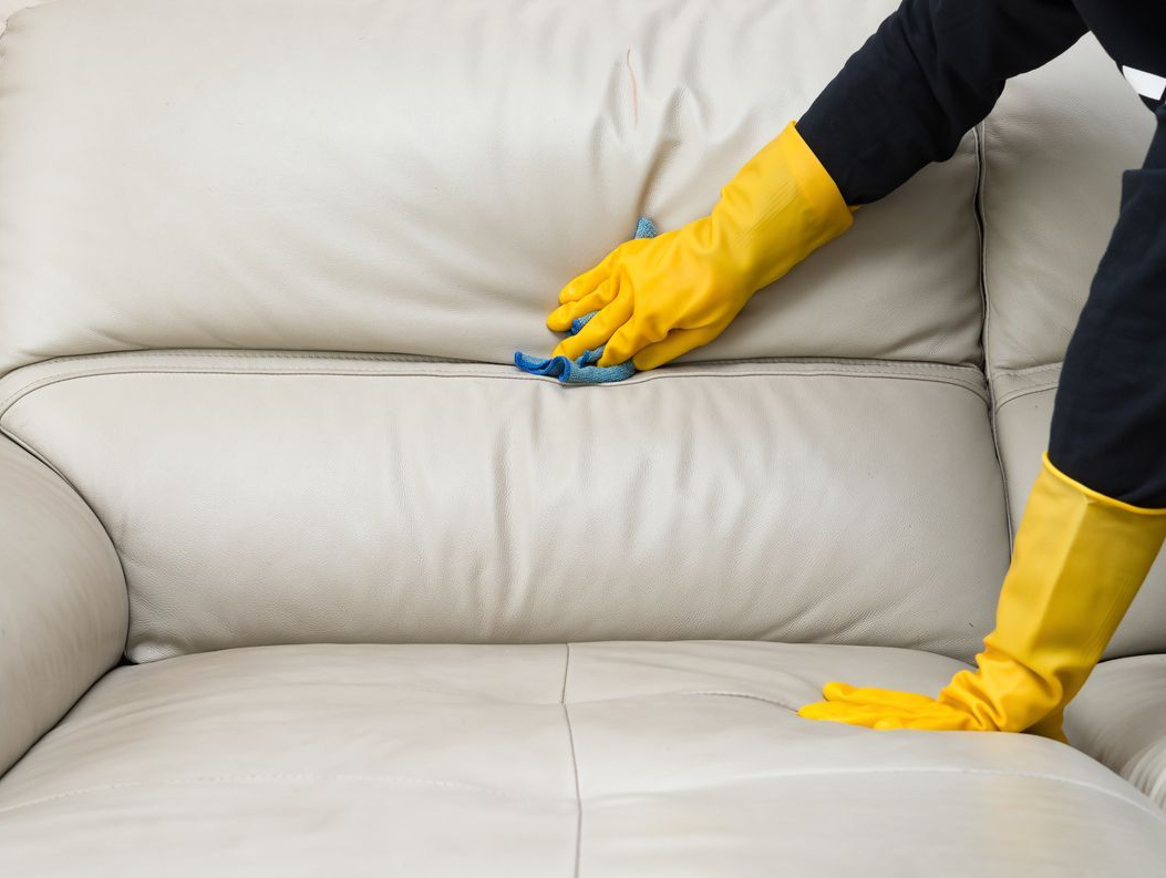 Pessoa com luvas amarelas limpando um sofá de couro branco com um pano azul.
