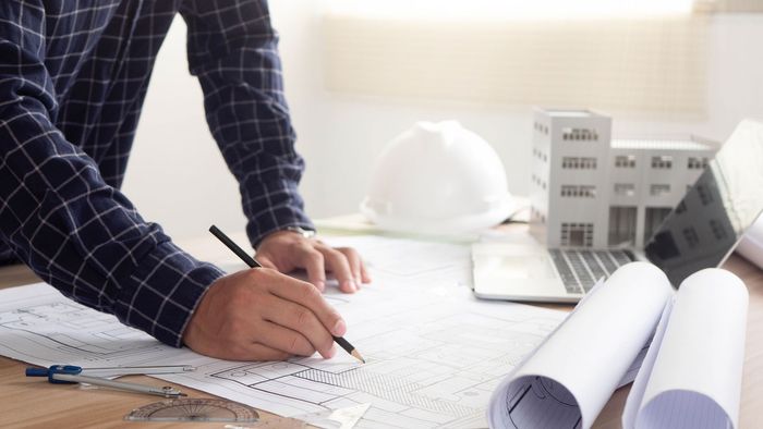Arquitecto sentado en su escritorio, dibujando planos con lápiz, con una maqueta y un ordenador portátil cerca.