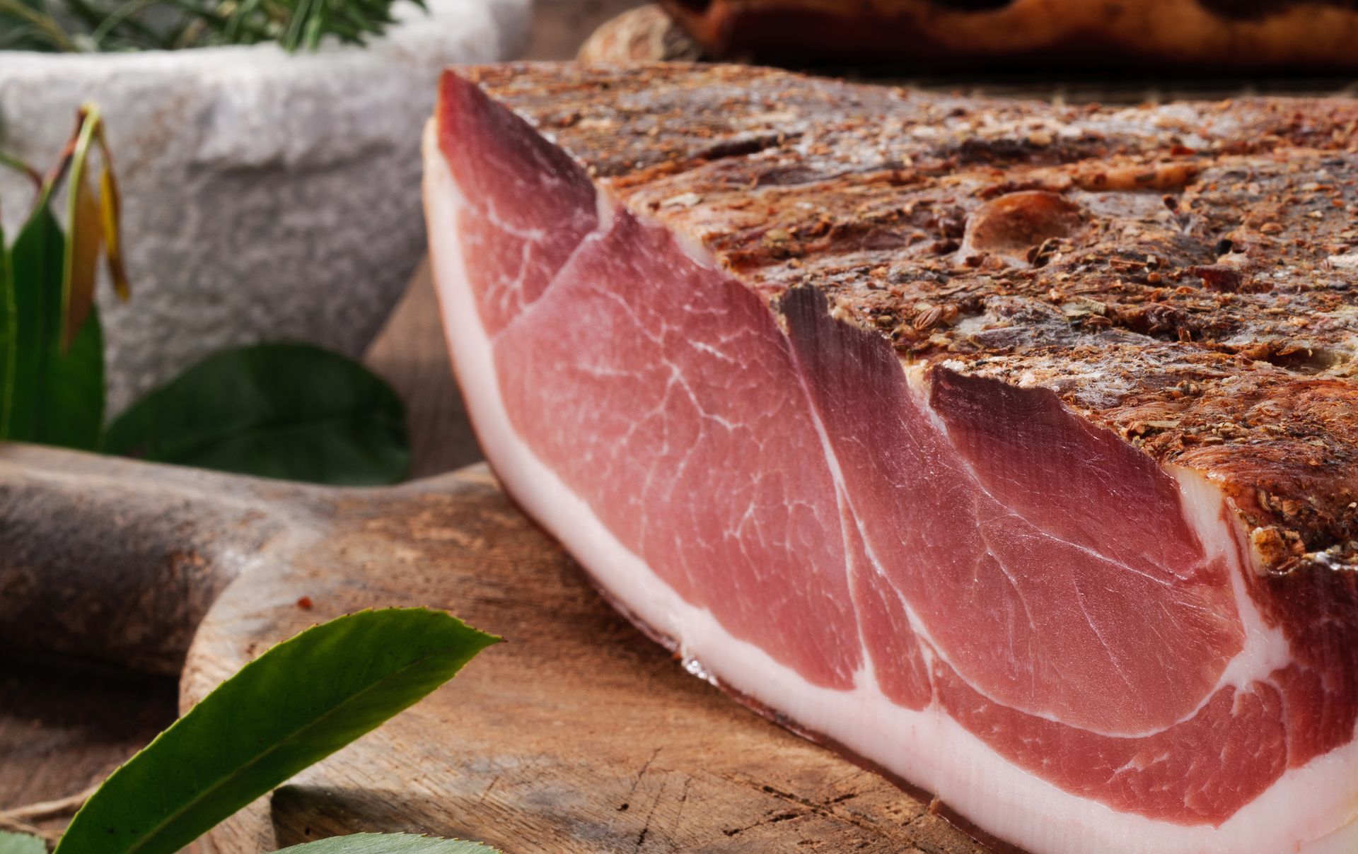 Gros plan sur une tranche de jambon cru, à la chair persillée de rose et à la croûte foncée, posée sur une planche en bois, à côté de feuilles vertes.