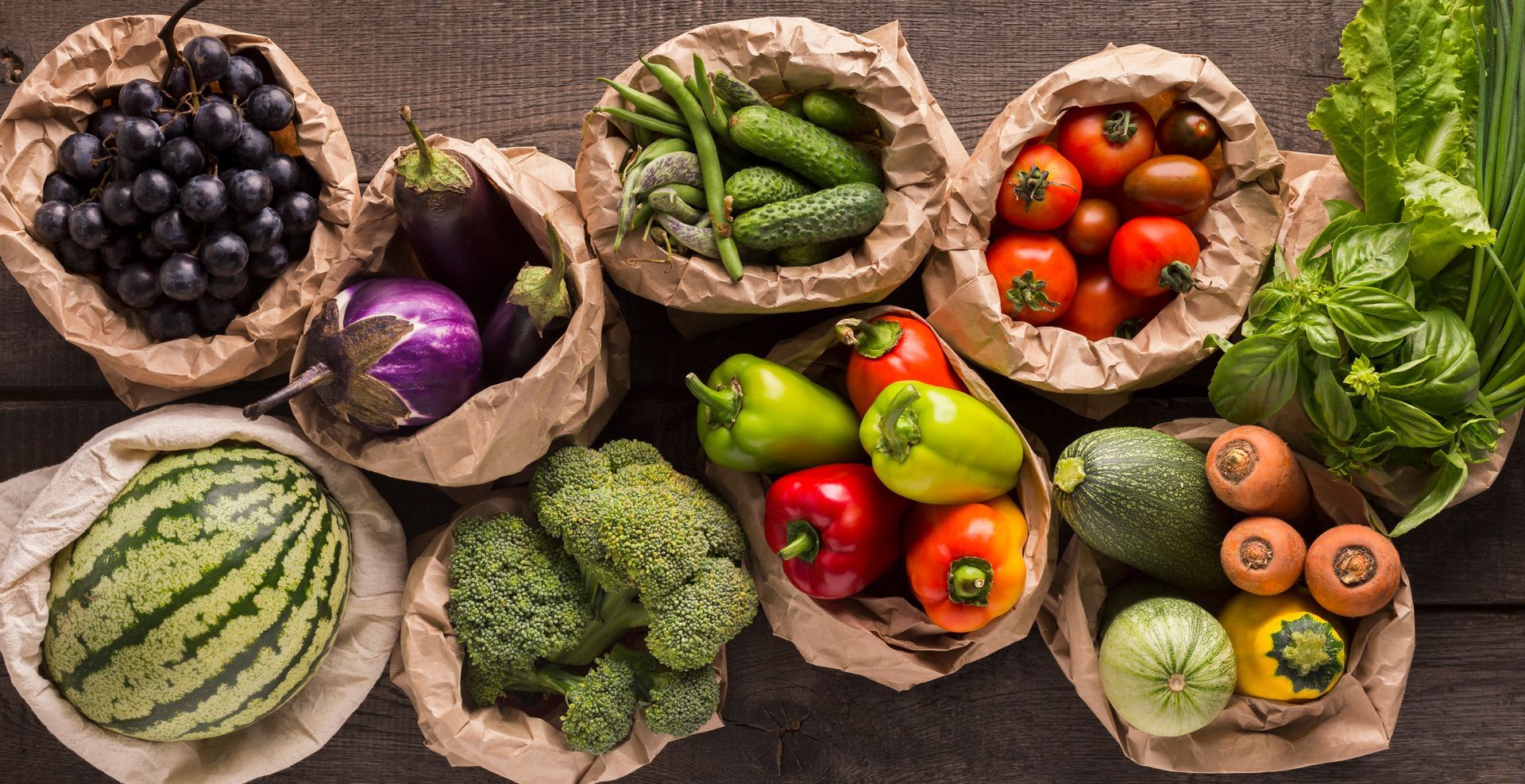 Fruits et légumes frais variés dans des sacs en papier sur une table rustique, notamment des baies, des poivrons, du brocoli et du melon.