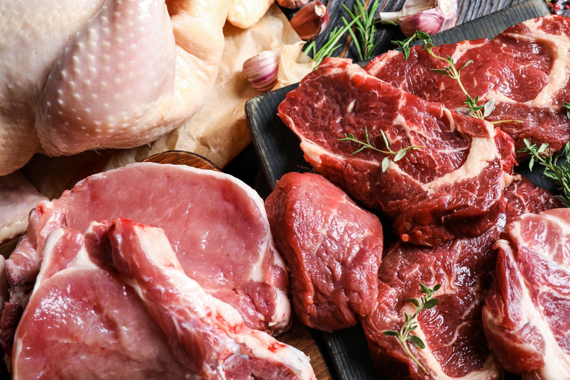 Des steaks de bœuf crus et un poulet entier sur un plateau, entourés d'herbes et d'ail.