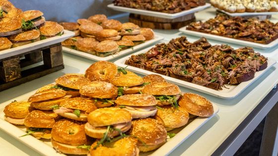 Plateau de sandwichs et viennoiseries assortis sur un buffet, avec d'autres plateaux de nourriture en arrière-plan.