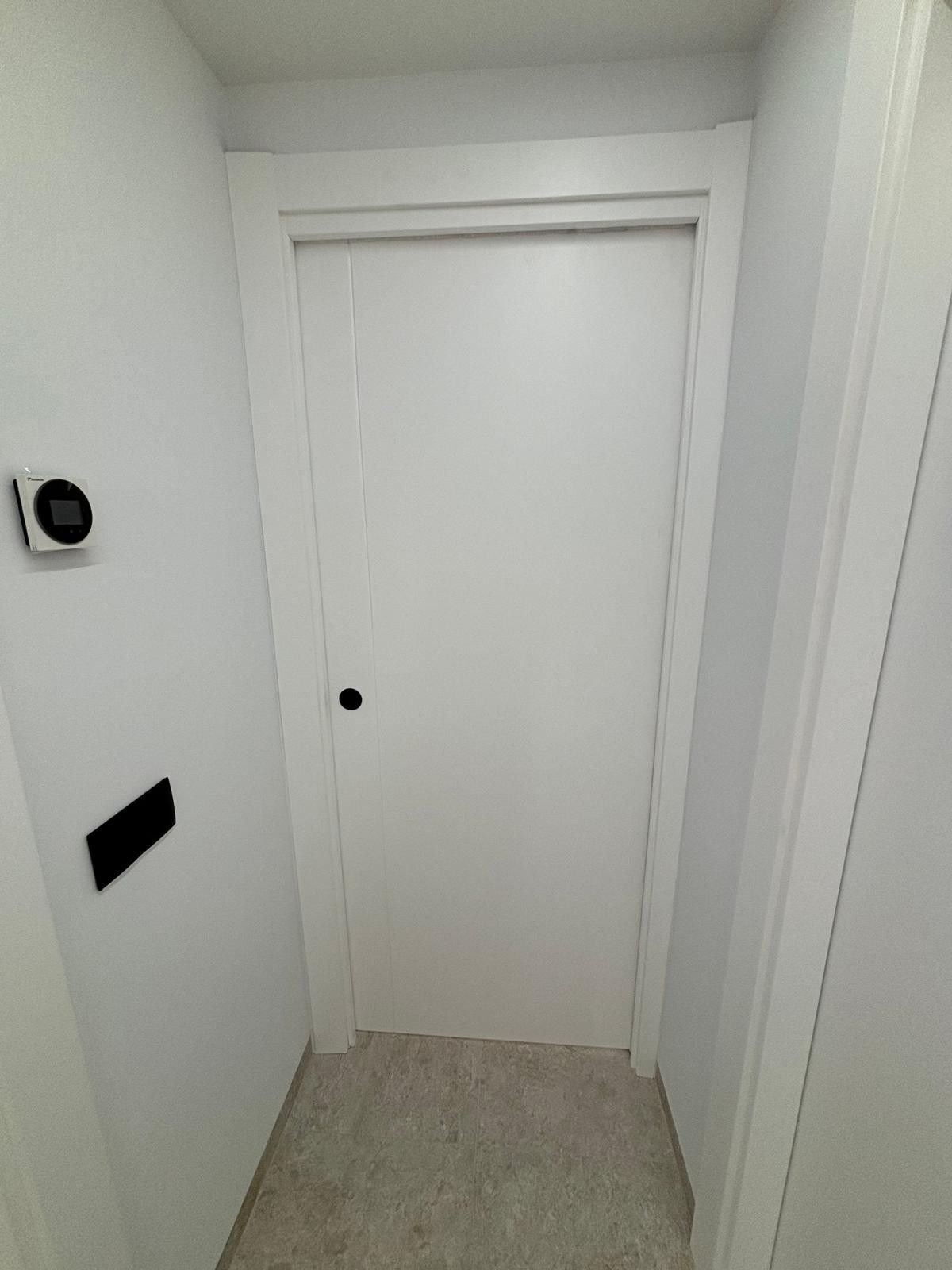 Puerta blanca en un pasillo con paredes blancas, manija de puerta negra y piso alfombrado.