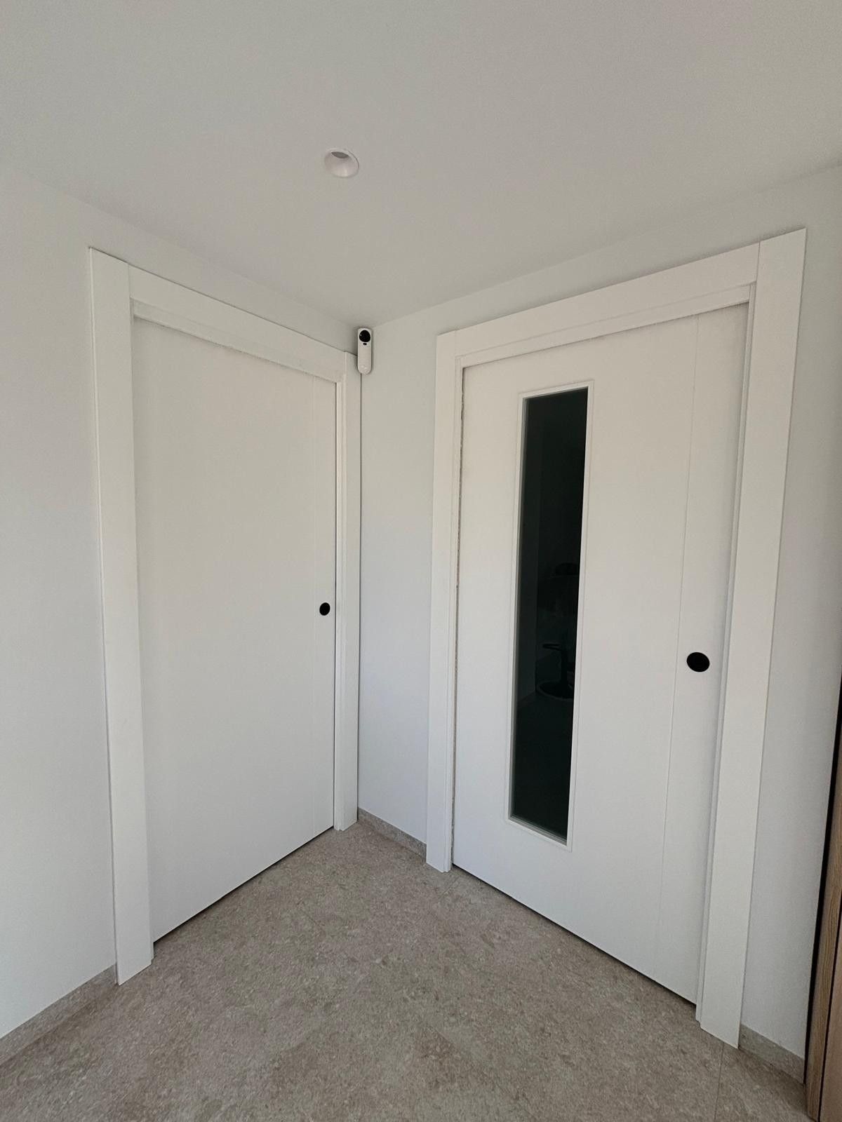 Dos puertas blancas en una esquina, una sólida y otra con un panel de vidrio vertical.