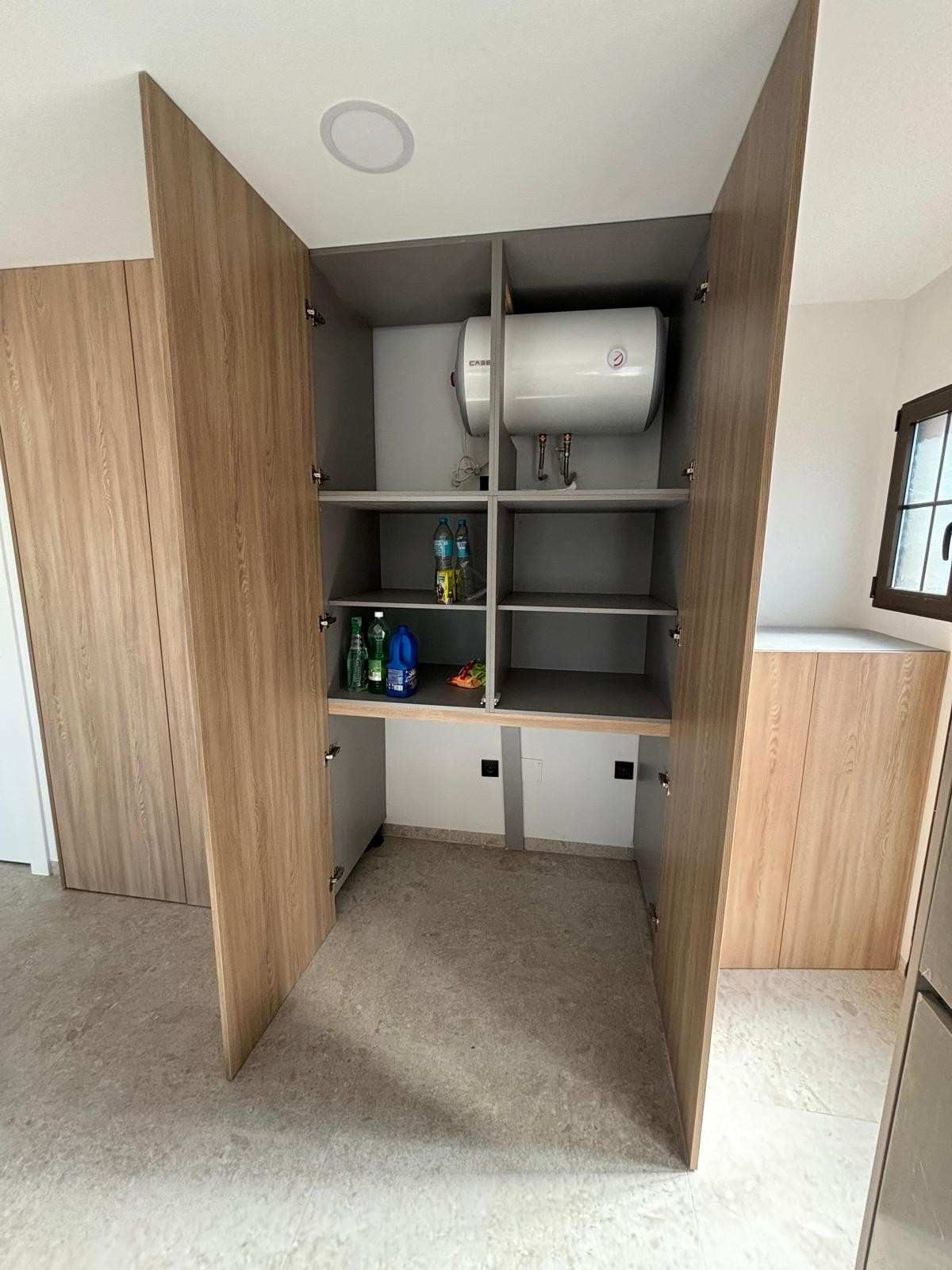 Mueble de madera con puertas abiertas, que alberga calentador de agua, estantes, artículos de limpieza y alfombra beige.