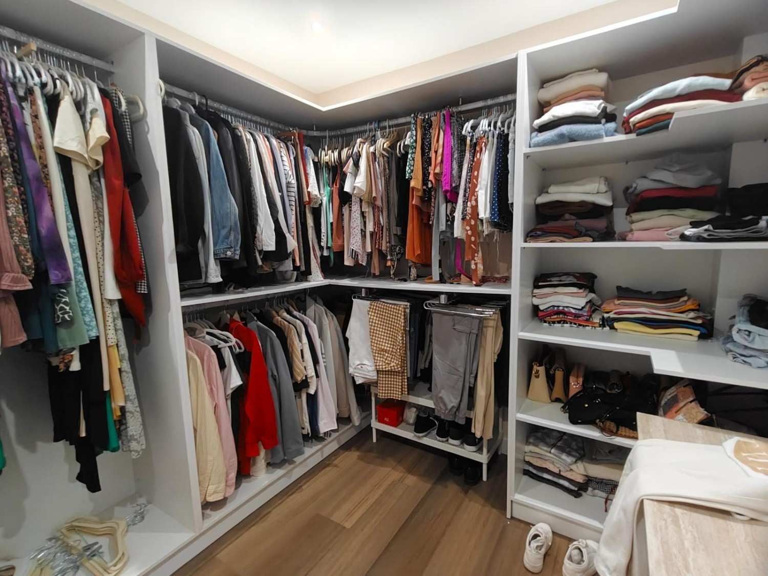 Vestidor con ropa en percheros y estantes; gabinetes blancos, piso de madera, iluminación brillante.