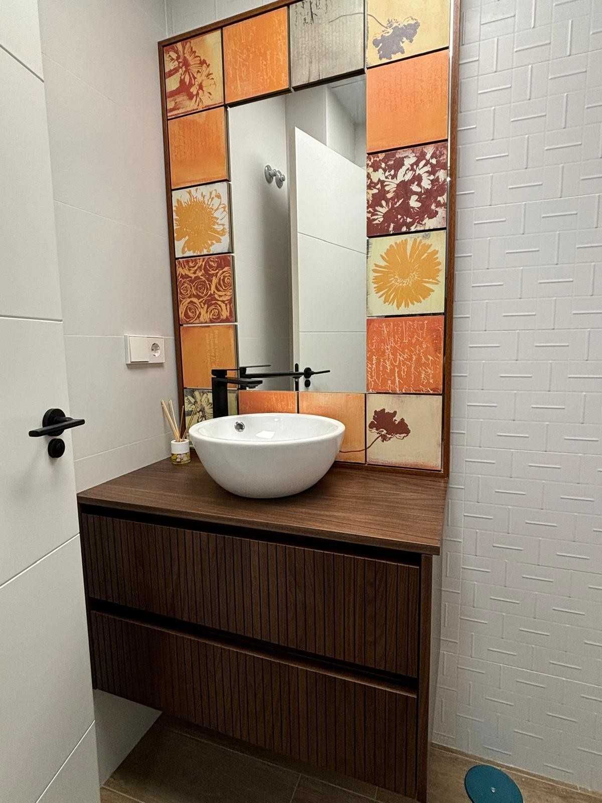 Mueble de baño con lavabo redondo blanco, mueble de madera y espejo enmarcado con azulejos de colores.