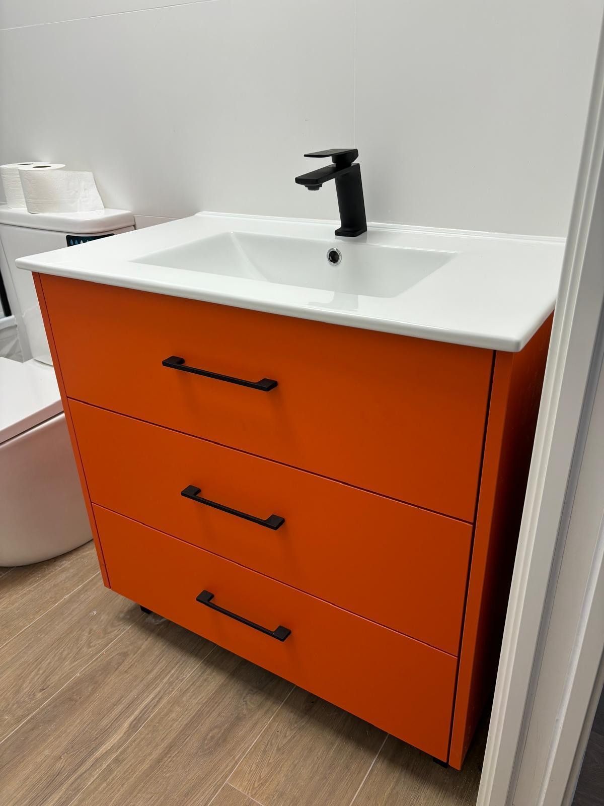 Mueble de baño color naranja con tres cajones, lavabo blanco y grifería negra.