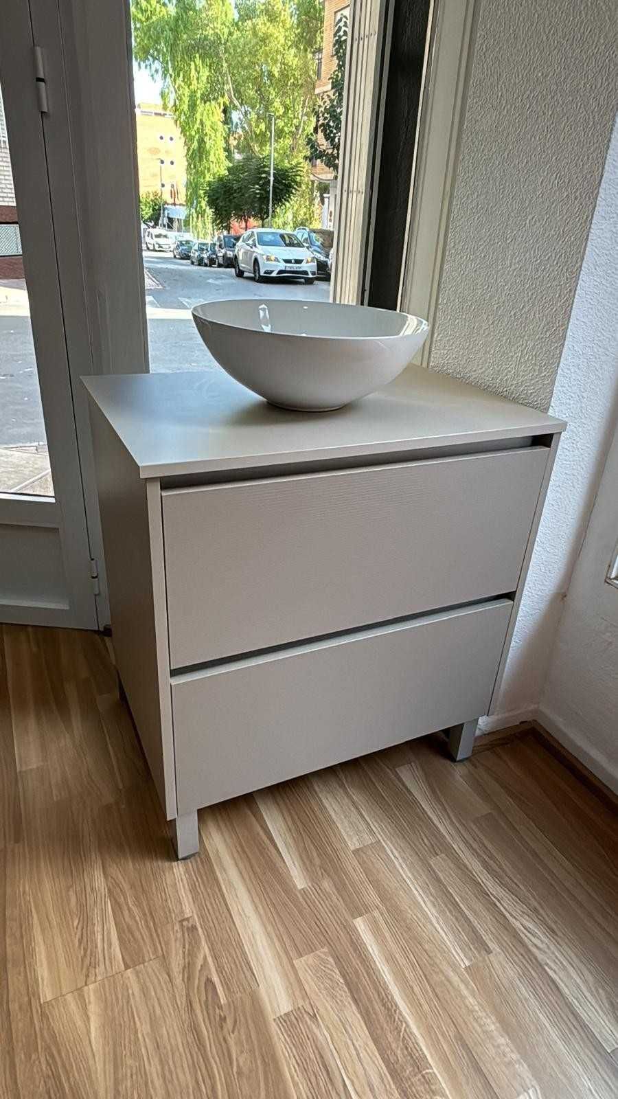 Armario gris claro con dos cajones y un cuenco blanco en la parte superior, situado cerca de una ventana.