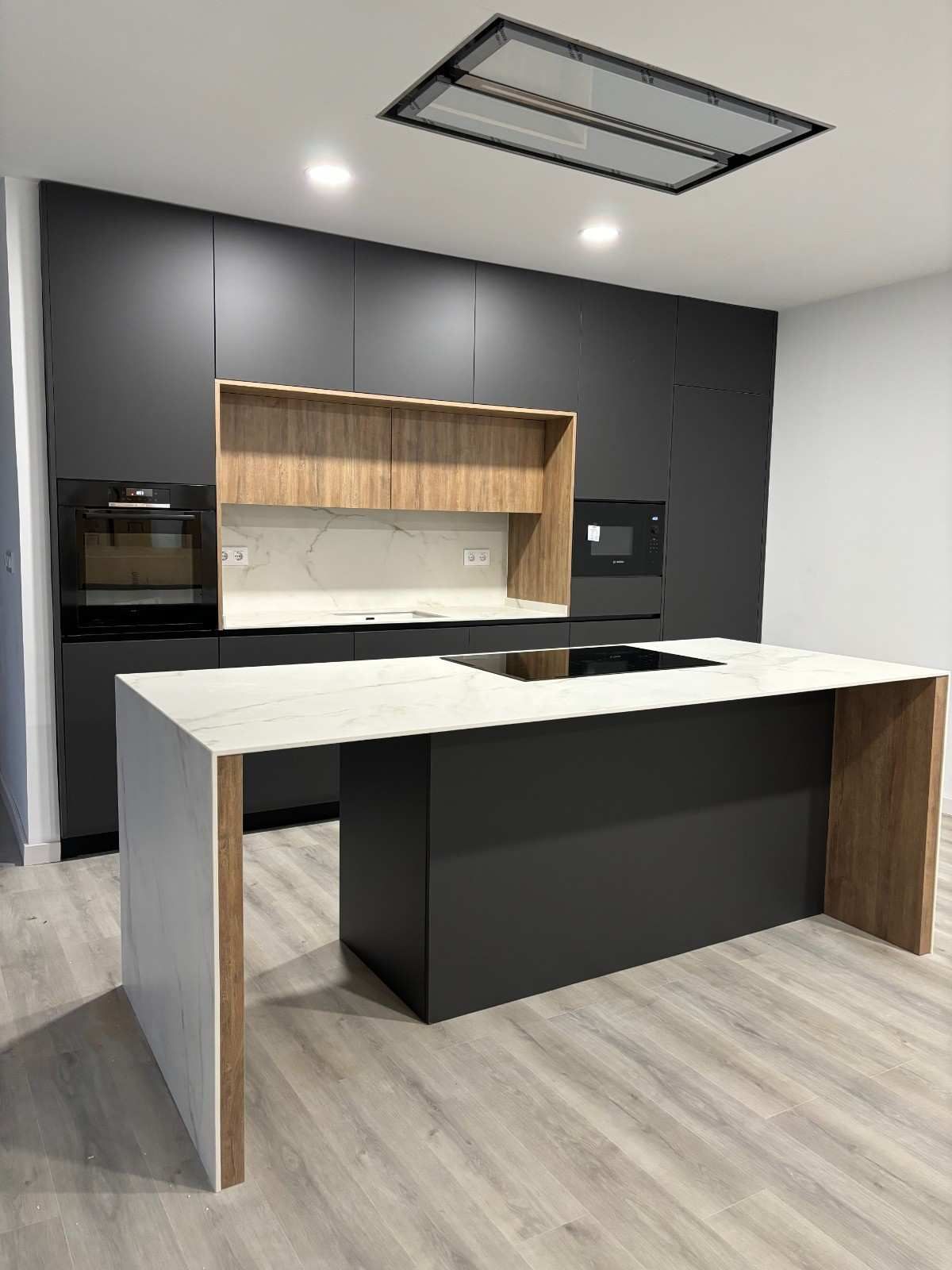 Cocina moderna con gabinetes de color gris oscuro, detalles de madera clara y una isla de encimera blanca.