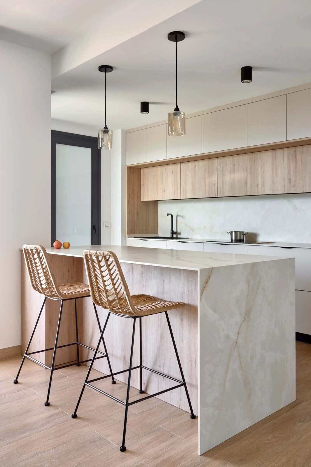 Cocina moderna con isla y gabinetes de madera clara, dos taburetes de bar de mimbre y luces colgantes.