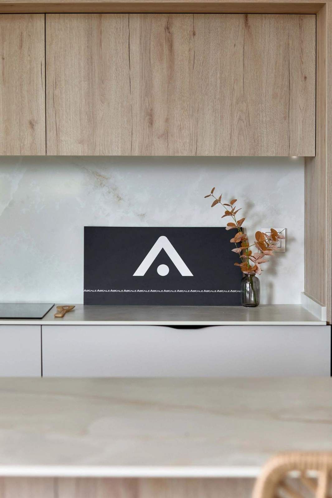 Cocina moderna con gabinetes de madera clara, encimera blanca, obras de arte negras con logotipo blanco y planta seca.