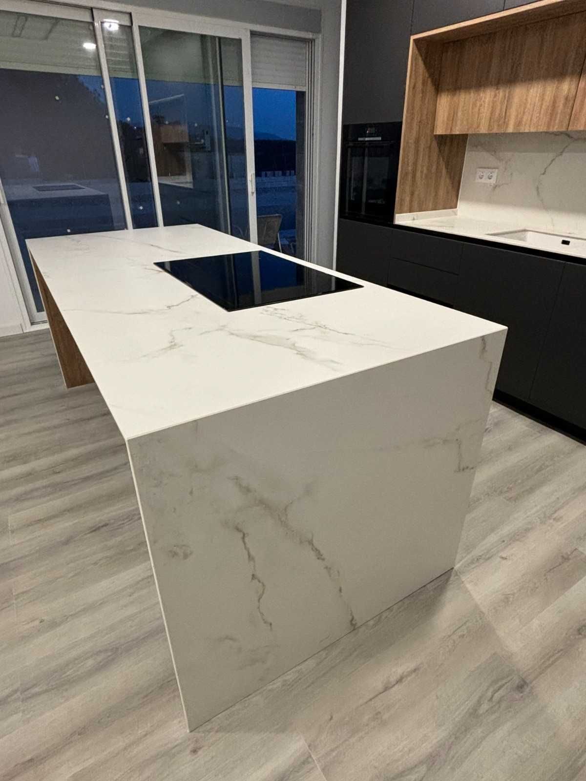 Isla de cocina de mármol blanco con placa de cocina integrada. Se ven muebles de madera clara y negros.