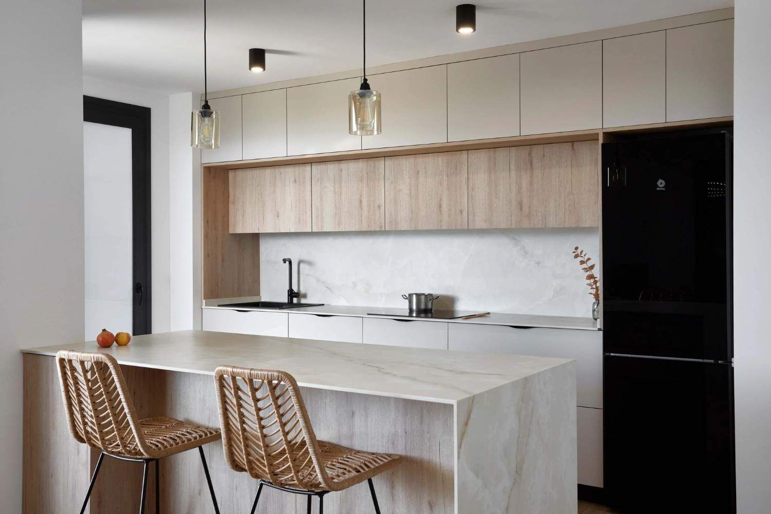 Cocina moderna con isla, gabinetes de madera clara, refrigerador negro y dos taburetes de bar de mimbre.