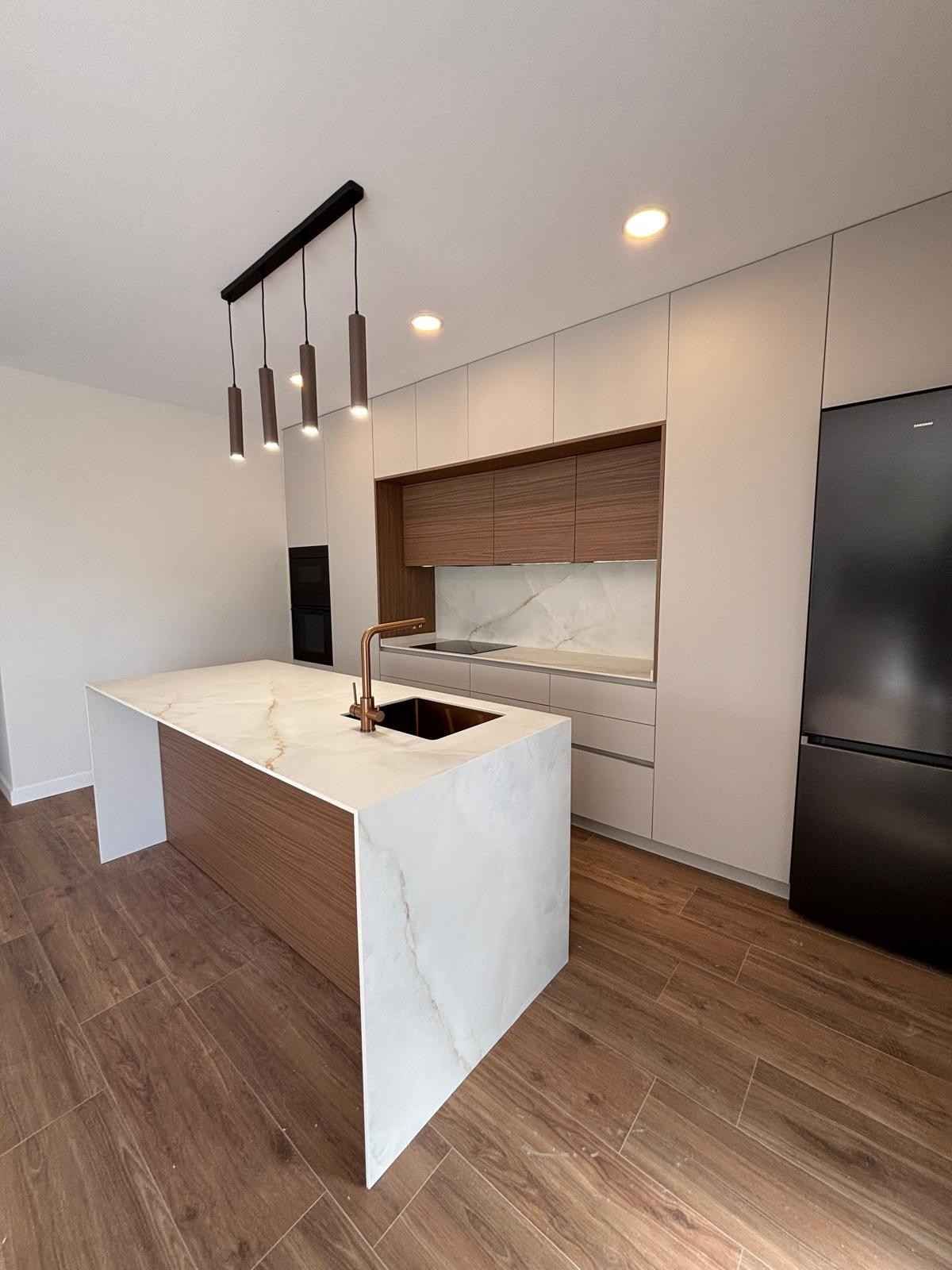 Cocina moderna con encimeras y gabinetes blancos, detalles de madera y refrigerador de acero inoxidable.