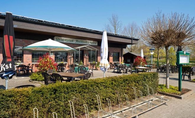 Restaurant „Buddeke's Aue“ mit Außenterrasse an einem sonnigen Tag. Braunes Gebäude, Tische, Sonnenschirme und grüne Hecken.