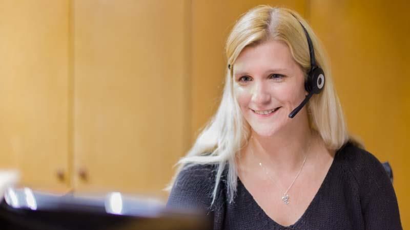 Eine Frau mit Headset sitzt lächelt an einem Schreibtisch.