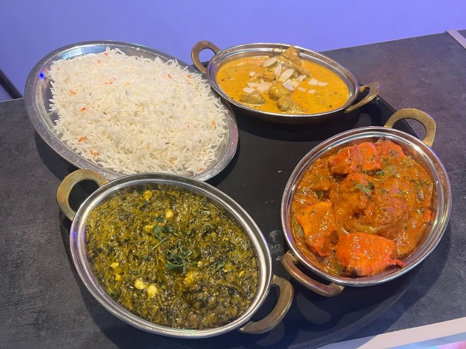 Quatre plats indiens présentés dans des bols de service en métal sur une table sombre : du riz, du curry, des légumes verts et un ragoût rouge.