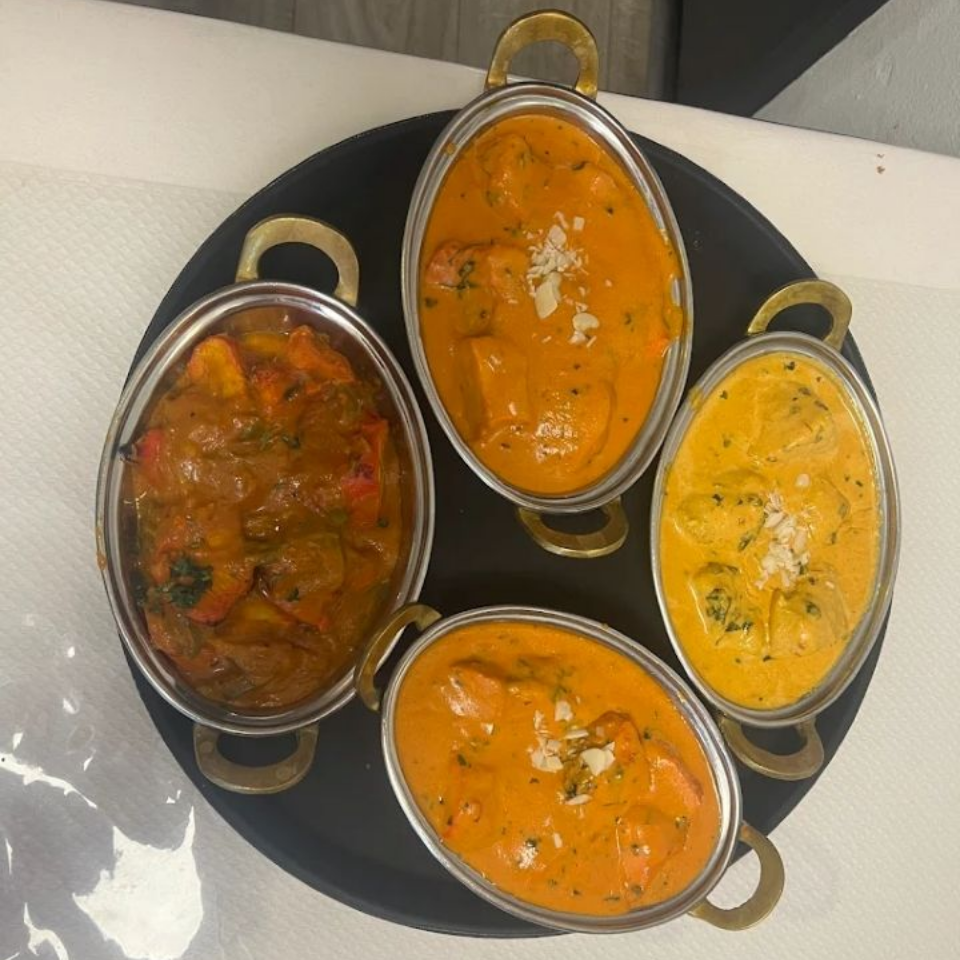 Quatre petits plats de curry dans des bols en inox, disposés sur un plateau noir, avec des sauces orange, jaune et rouge.