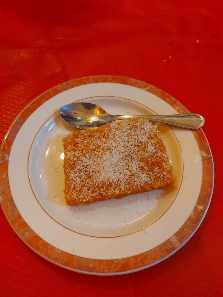Une part de gâteau saupoudrée de noix de coco, posée dans une assiette blanche et or, avec une cuillère sur une nappe rouge.