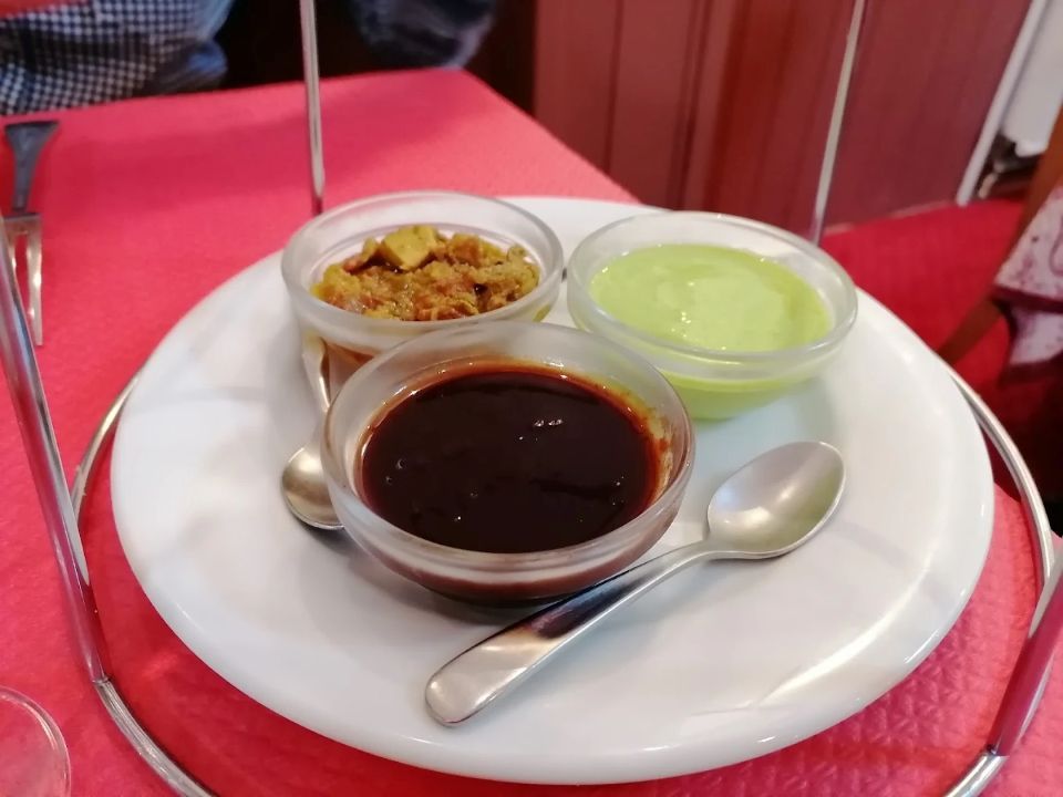 Trois coupes de dessert sur une assiette blanche avec des cuillères, contenant une sauce brune et un pudding vert, sur une nappe rouge