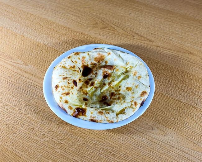 Assiette blanche avec un naan sur une table en bois, légèrement dorée et garnie de verdure.
