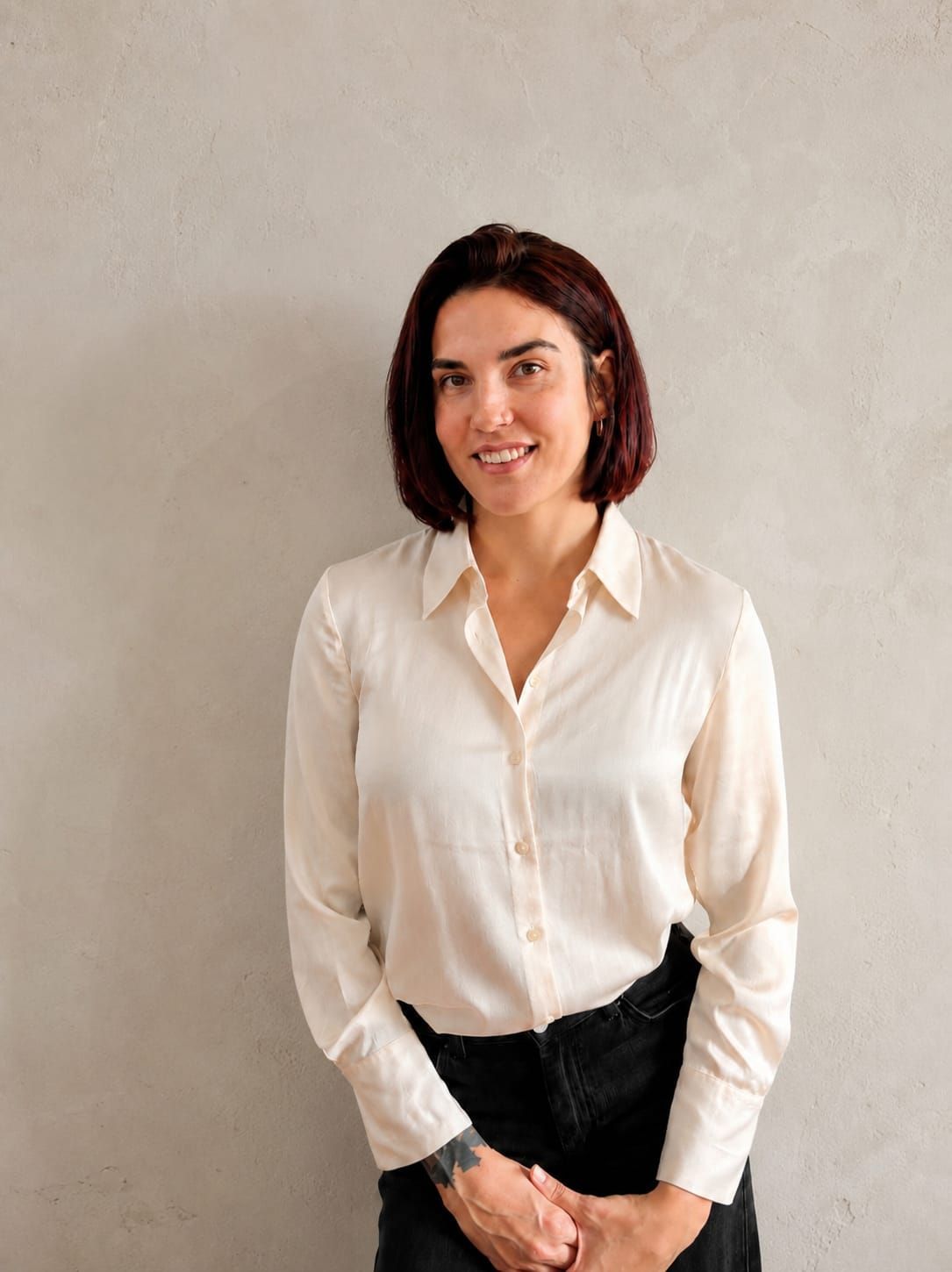 Mujer con blusa blanca y pantalones negros de pie contra una pared lisa, sonriendo a la cámara.