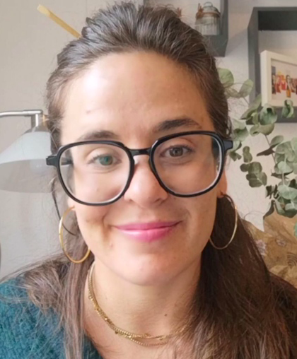 Persona sonriente con gafas y pendientes de aro, en el interior de una casa con una planta y una lámpara al fondo.