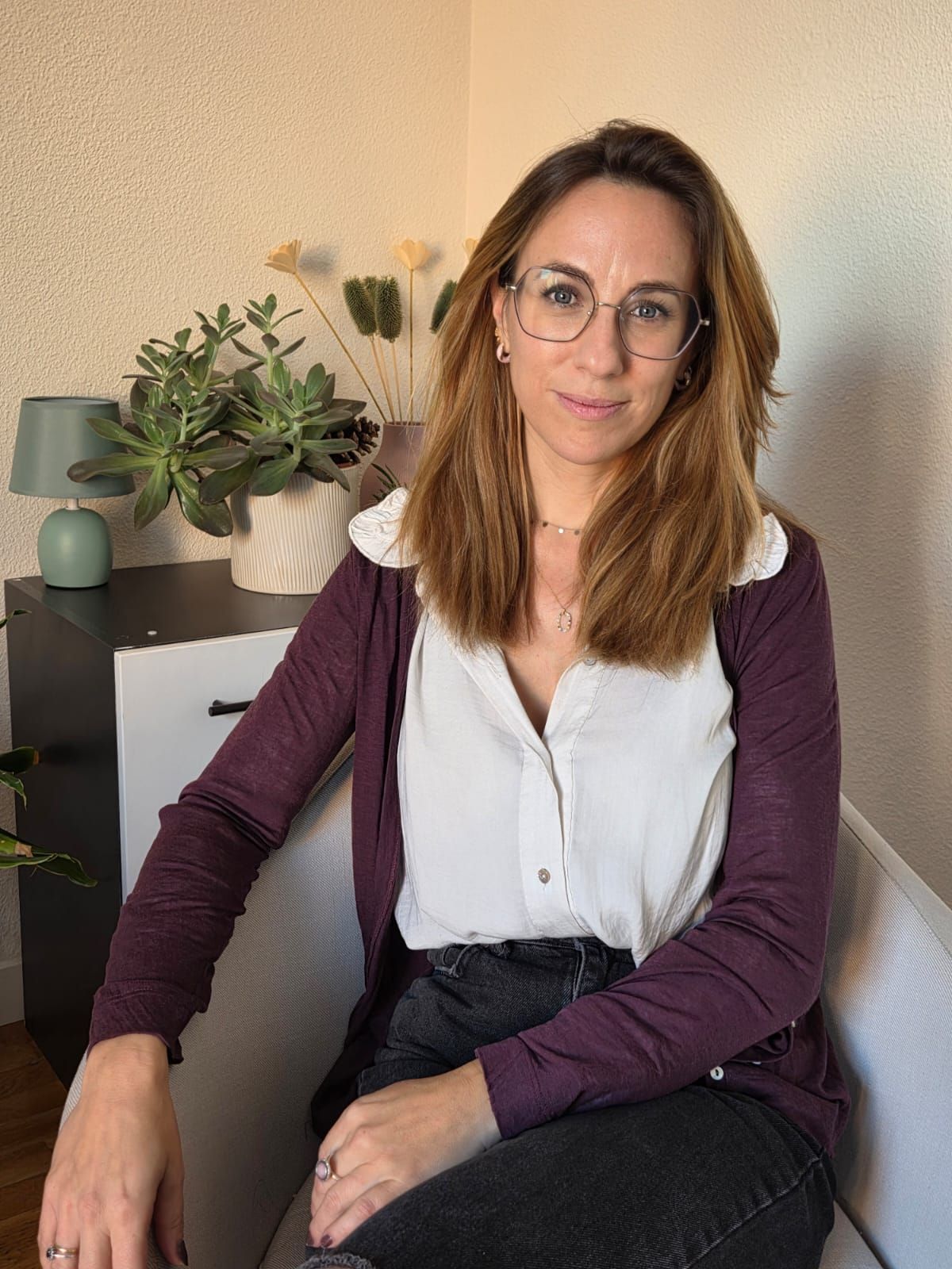 Mujer sentada en el interior de una casa, junto a unas plantas, con gafas, blusa blanca y cárdigan morado.