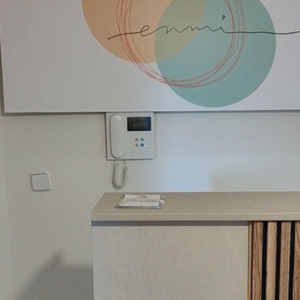 Intercomunicador montado en la pared sobre un mueble de madera clara, con arte abstracto en tonos pastel en la pared y un auricular colgante.