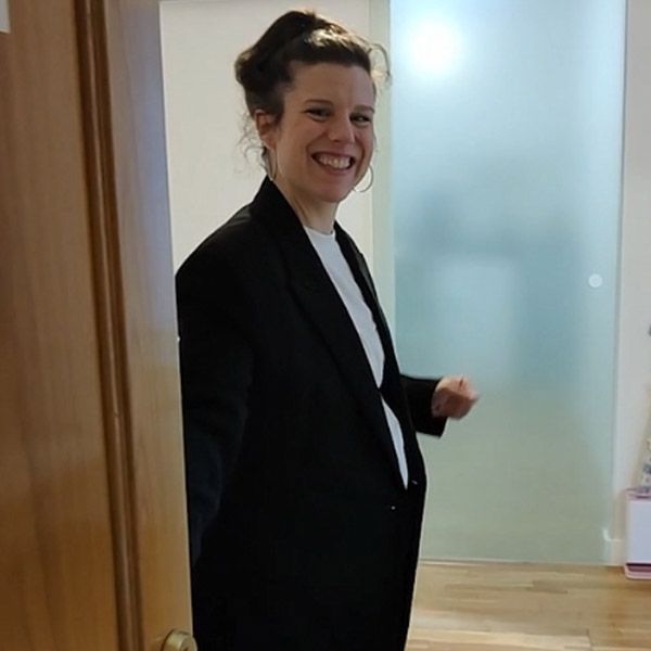 Una persona con un blazer negro sonríe en el umbral de una puerta, junto a una pared de vidrio esmerilado.