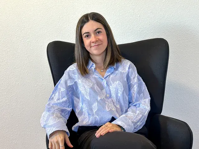 Una mujer con una blusa azul claro está sentada en una silla negra, sonriendo, en el interior.