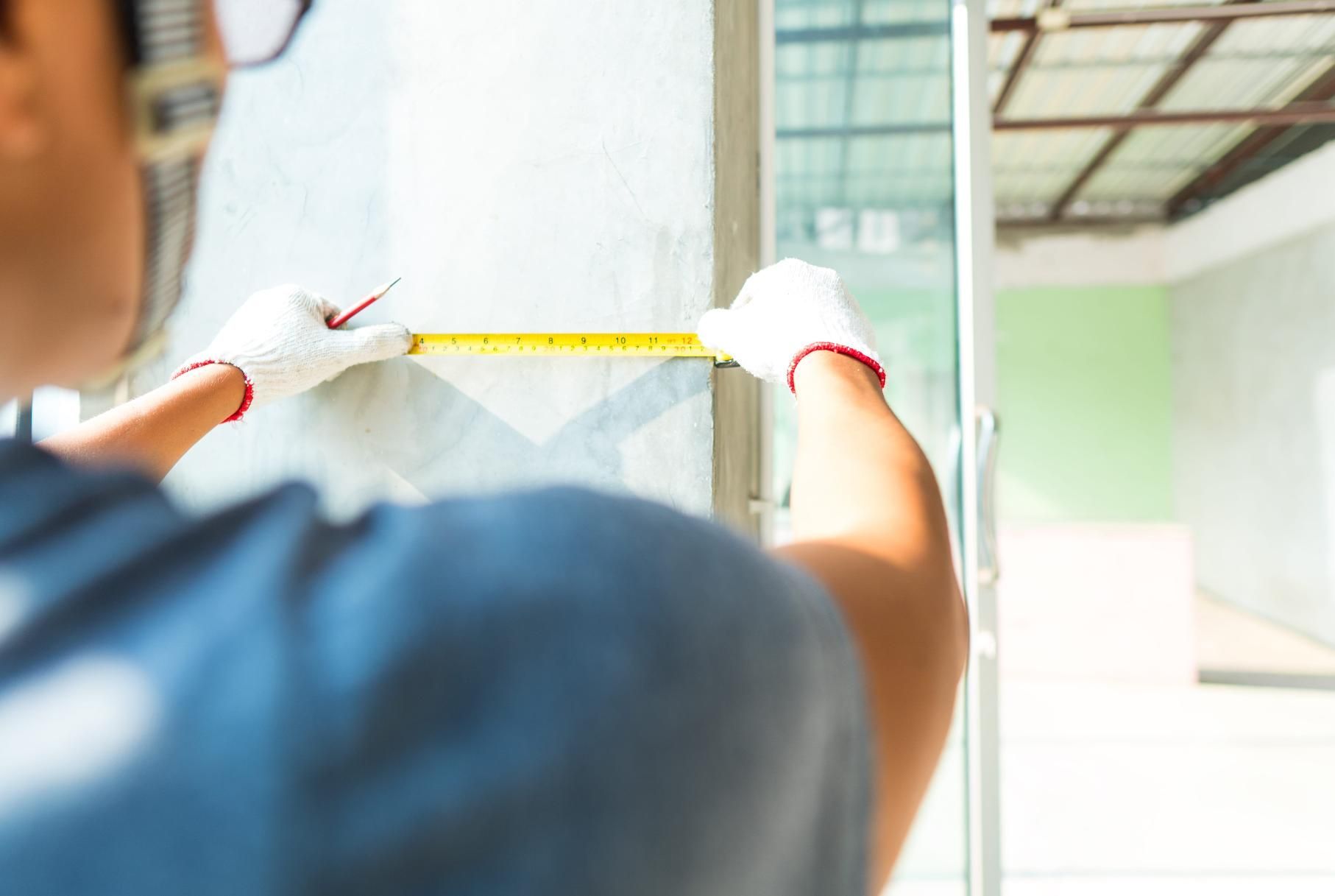 Un obrero de la construcción, con casco y guantes blancos, utiliza una cinta métrica amarilla en el marco de una pared.