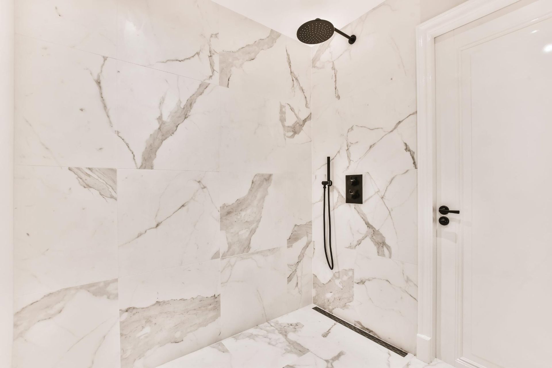 Una ducha a ras de suelo con azulejos que imitan el mármol blanco, un cabezal de ducha negro tipo lluvia