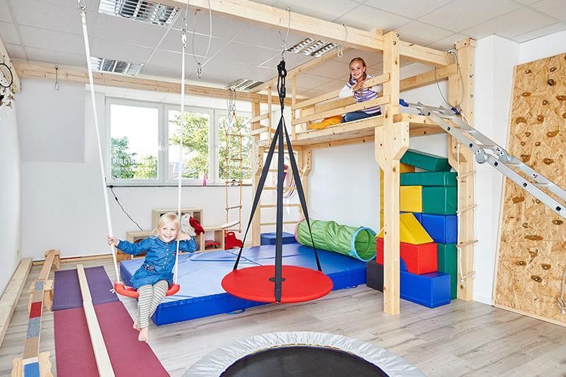 Zwei Kinder spielen in einem Raum mit einem Trampolin und Schaukeln.