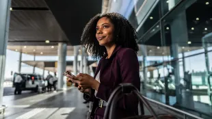 Mujer con un traje morado en un aeropuerto, sosteniendo un teléfono, mirando hacia arriba y llevando un bolso.