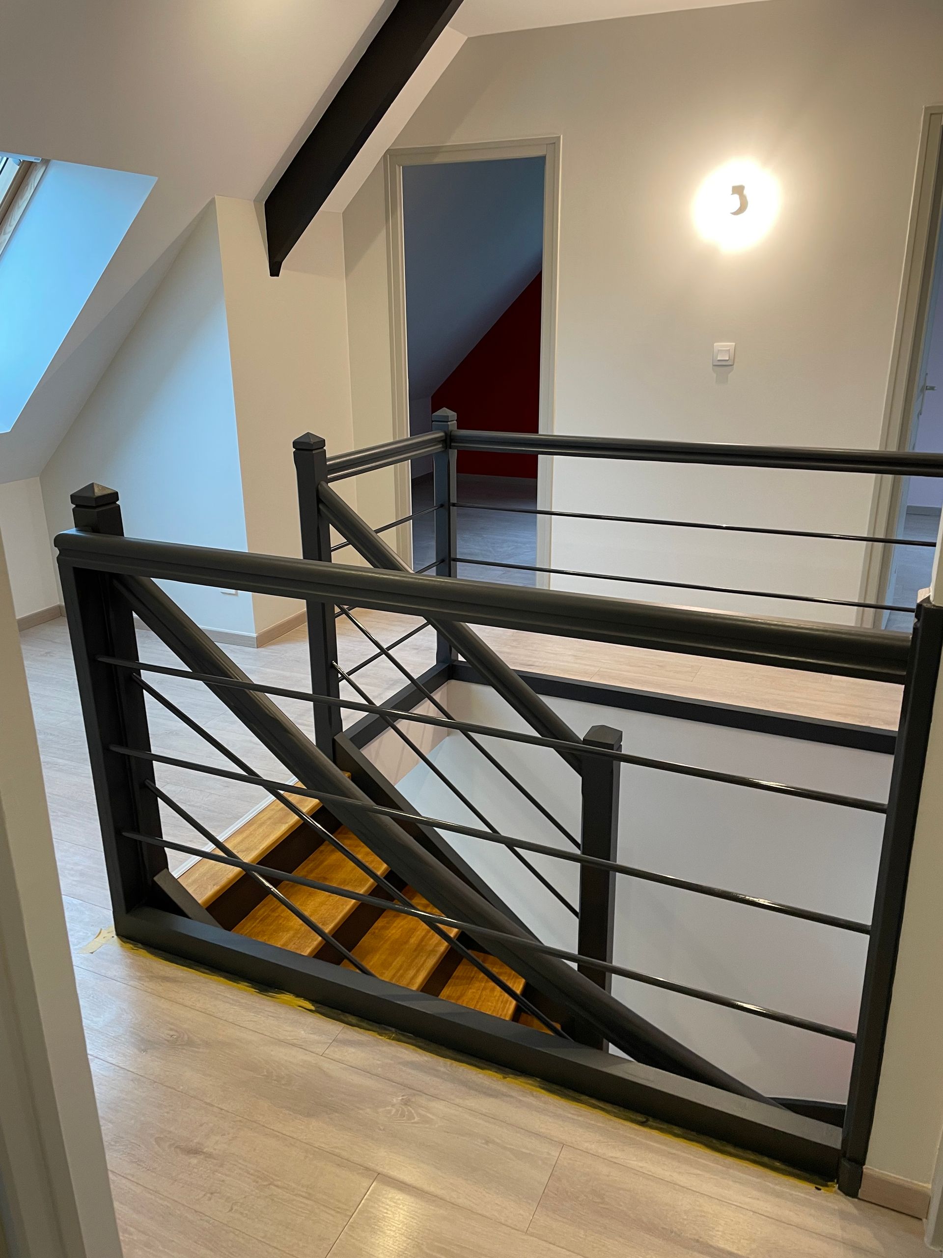 Cage d'escalier avec rampe en métal et marches, vue depuis le palier de l'étage supérieur.