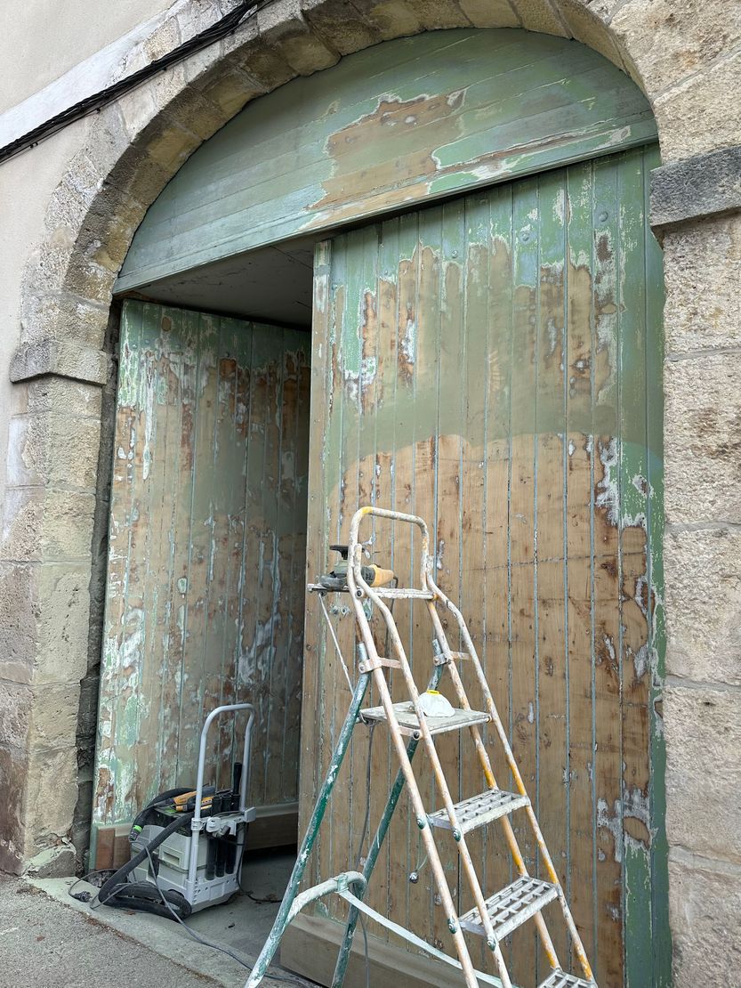 Une échelle appuyée contre une porte cochère en bois usée par le temps, dans une arche en pierre.