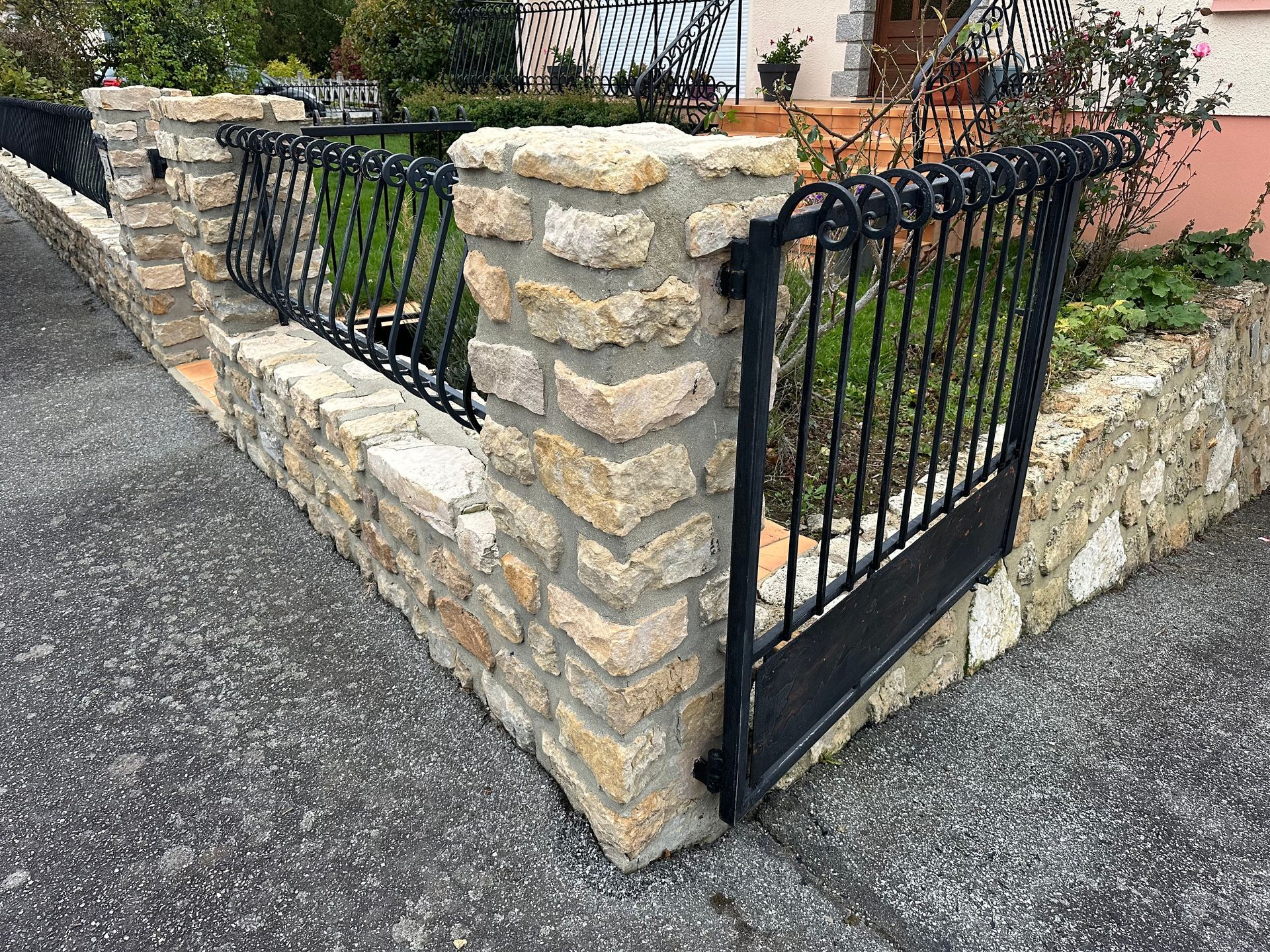 Mur de jardin en pierre avec portail et clôture en métal le long d'une allée pavée.