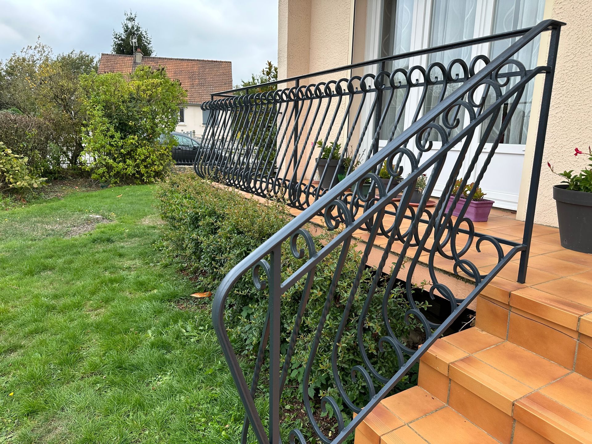 Rambarde d'escalier en métal à côté de marches carrelées menant à une maison, avec un jardin et une pelouse à proximité.